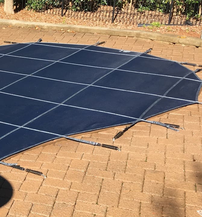 A dark blue safety cover stretched across a pool, anchored to a brick paver deck