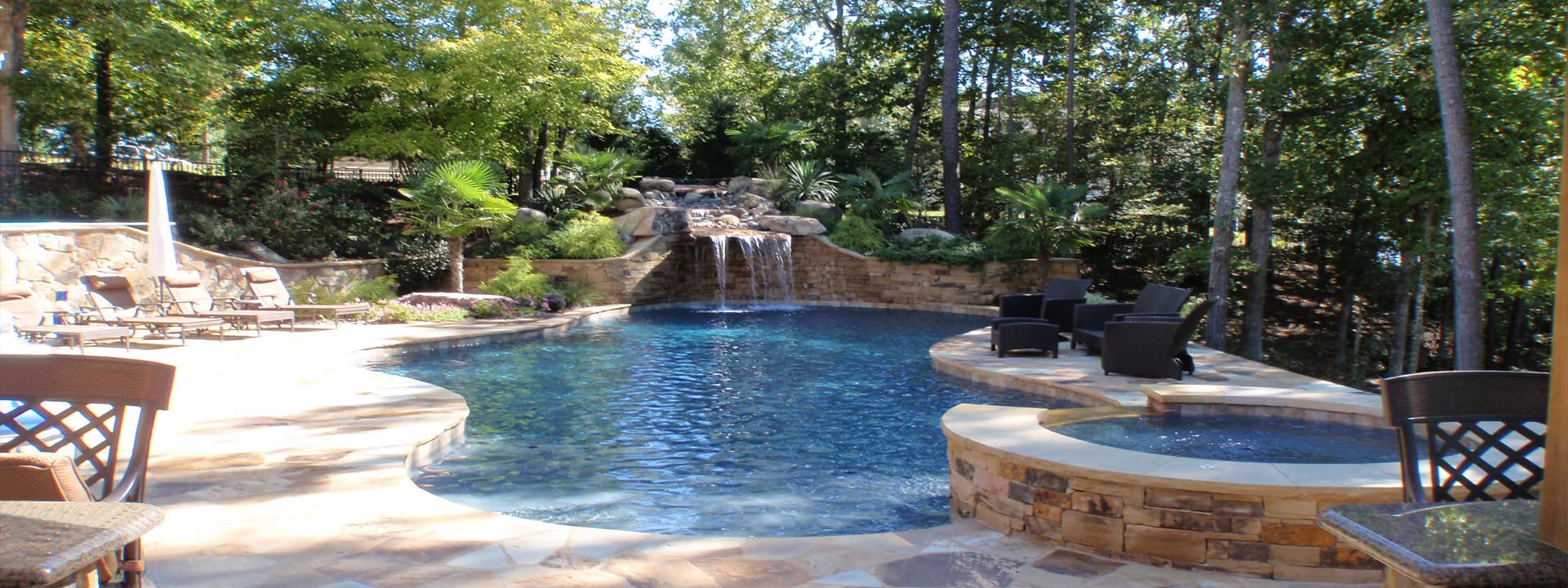 A backyard pool with a small waterfall, a stone-edged spa and patio seating among tall trees
